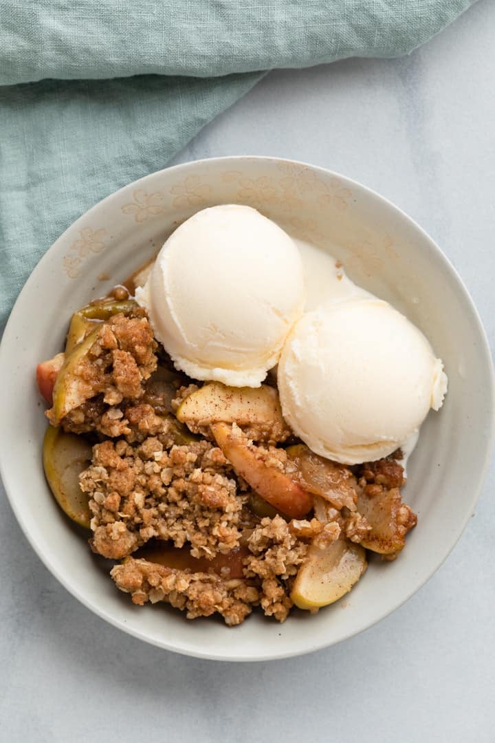 overhead view of apple crisp in grey bowl with 2 scoops of vanilla ice cream