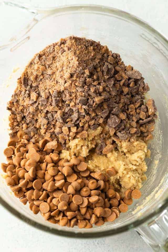 butterscotch chips and chocolate toffee pieces added to cookie dough in a glass bowl