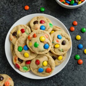 M&M cookies on a white plate with a bowl of m&m's next to it and a couple cookies scattered about on the table.