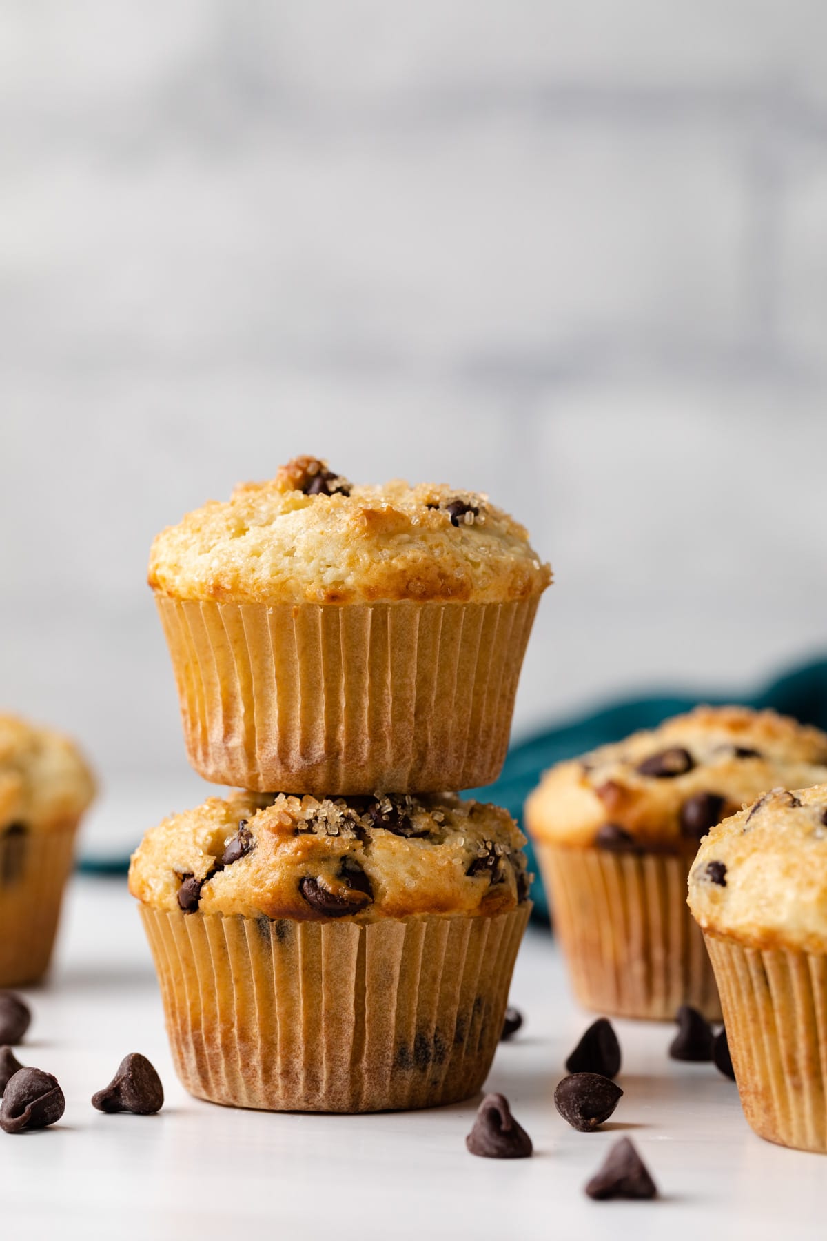 two chocolate chip muffins stacked with more muffins and chocolate chips scattered around