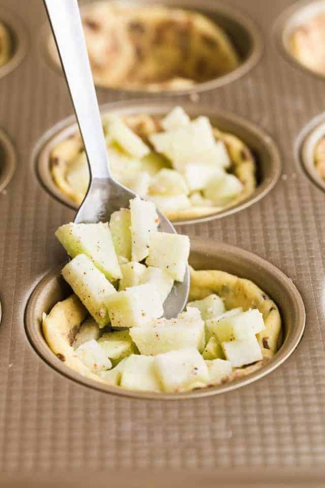 Cinnamon roll cups in a muffin tin being filled with chunks of apple.