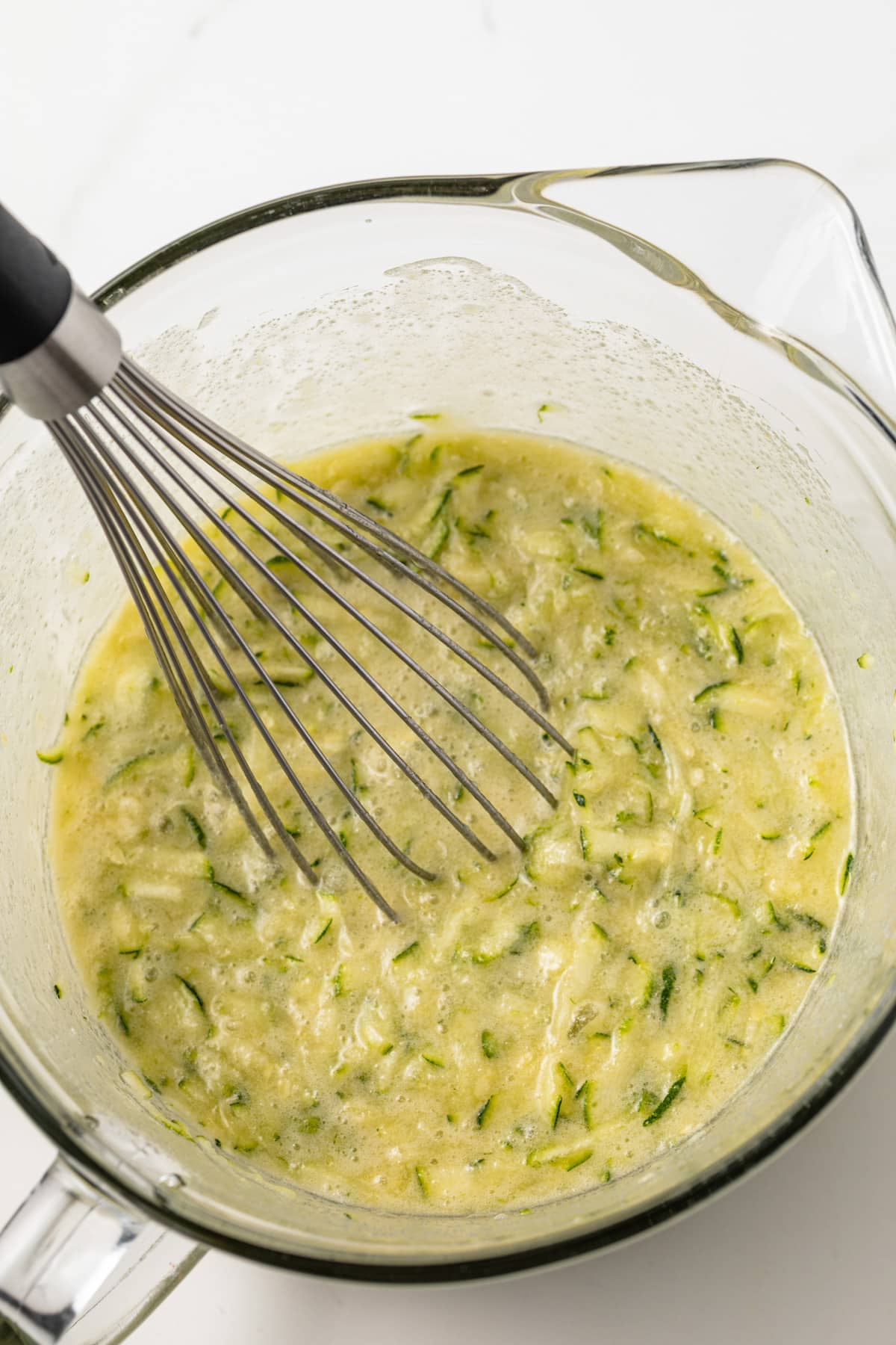 Shredded zucchini mixed into wet ingredients in a glass bowl.
