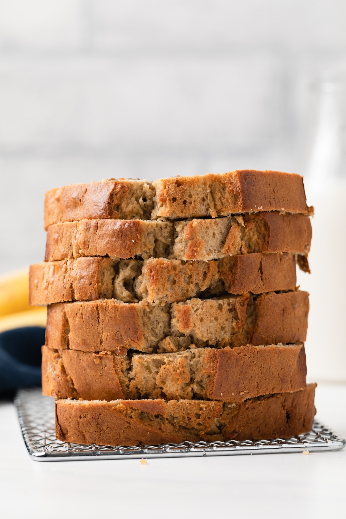 side view of six slices of easy banana bread stacked on wire rack with bananas and jar of milk in background
