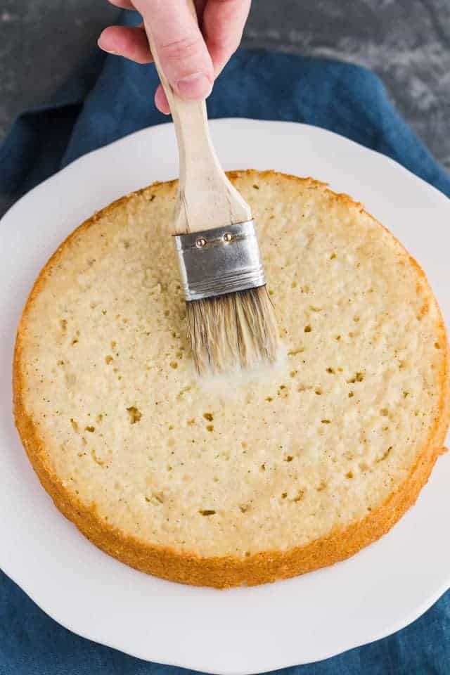 Soaking syrup being brushed over a layer of eggnog cake on a white cake stand.