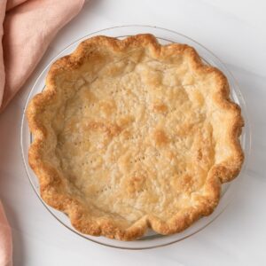 overhead of blind baked pie crust in glass pan with peach colored napkin
