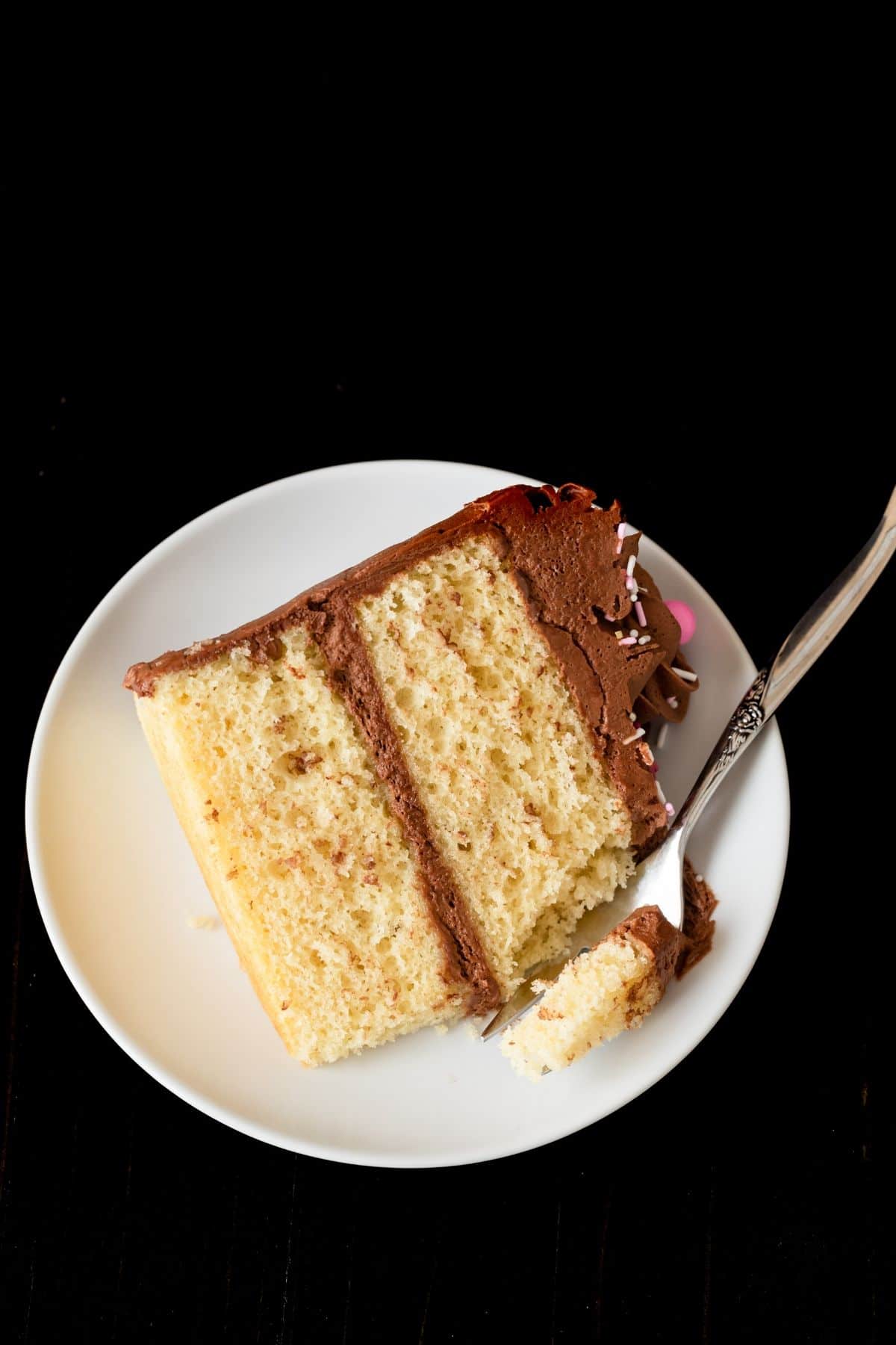 Slice of birthday cake on a white plate with a fork taking a bite out.