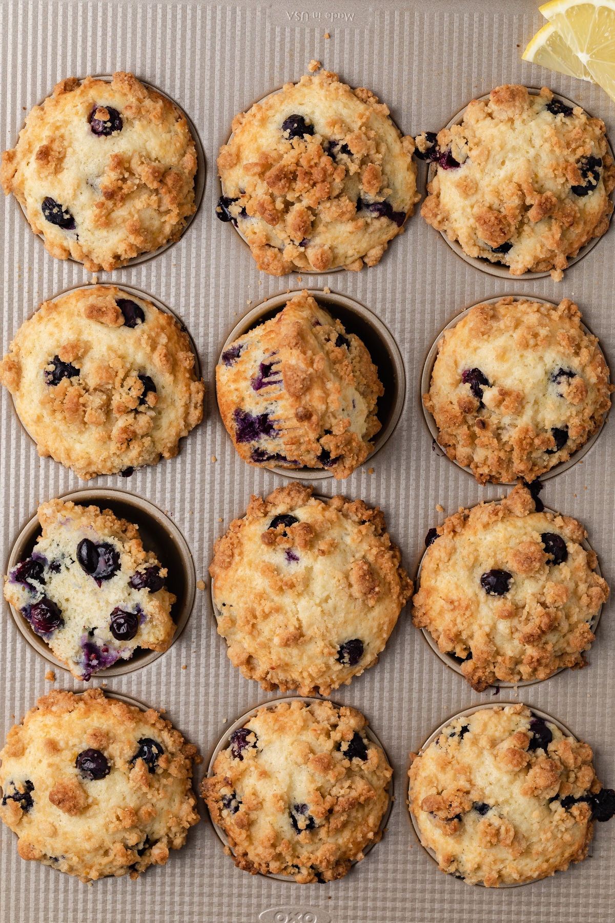 overhead of lemon blueberry muffins in a muffin pan with one sliced in half so the inside is visible