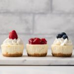 three mini cheesecakes topped with whipped cream and fruit on a marble platter