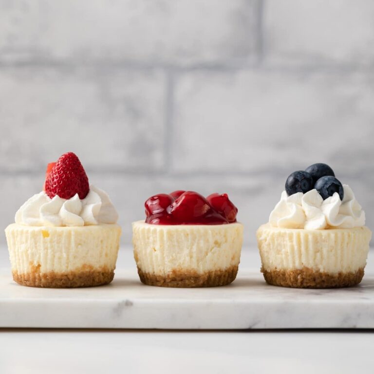 three mini cheesecakes topped with whipped cream and fruit on a marble platter