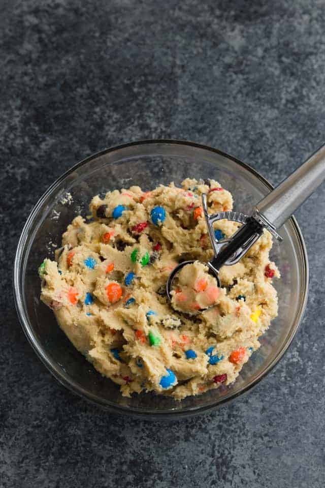 M&M cooking dough in a glass bowl with a cookie scoop portioning the dough.