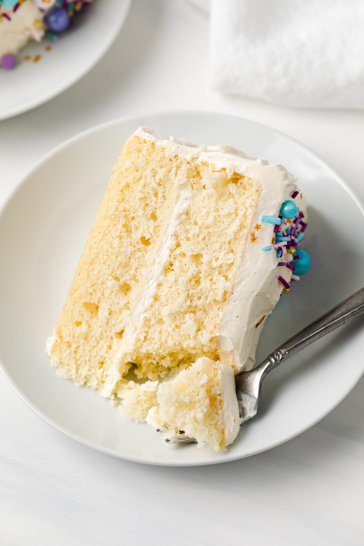 angled view of a slice of moist vanilla cake on white plate with a fork taking out a bite