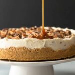no bake snickers cheesecake with caramel drizzle on a white cake stand