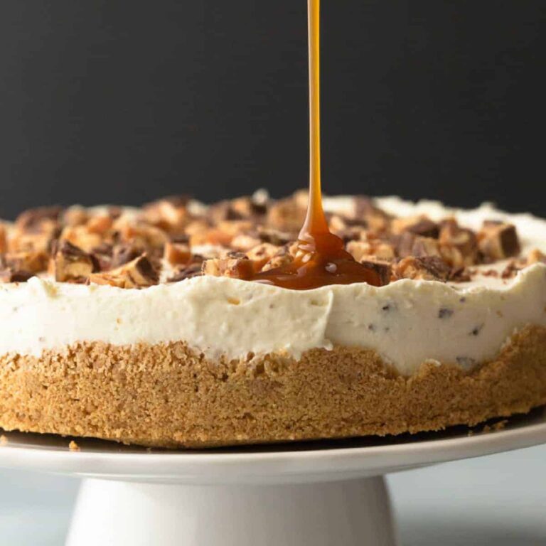 no bake snickers cheesecake with caramel drizzle on a white cake stand