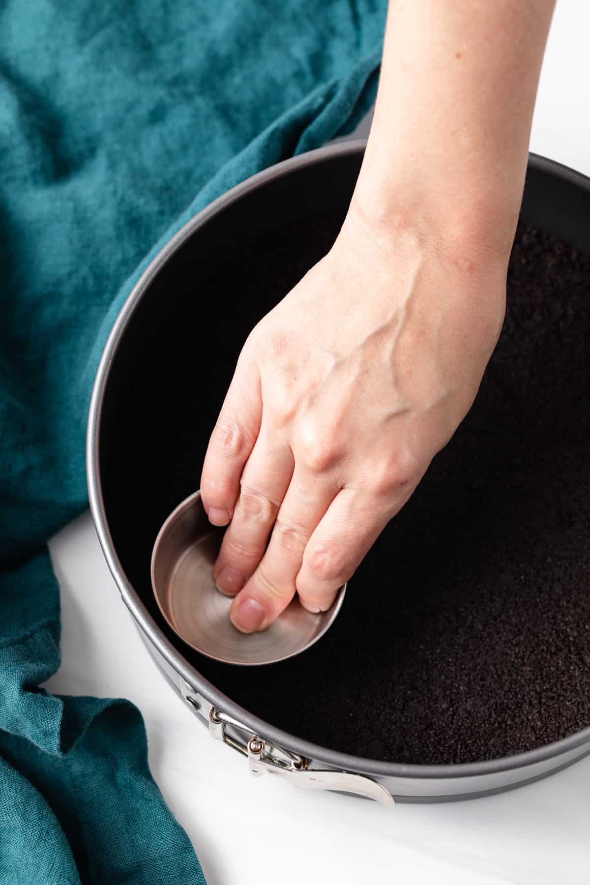 hand pressing oreo crumbs into cheesecake pan with a measuring cup