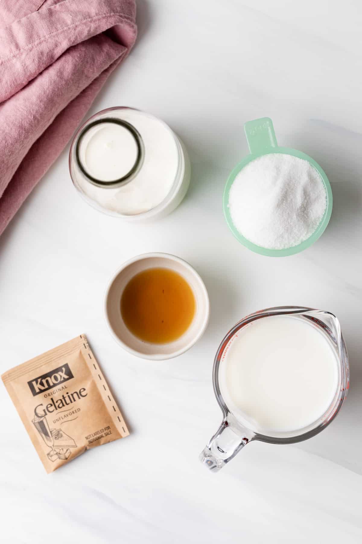 Milk in a glass measuring cup, cream in a glass jar, sugar in a jade measuring cup, vanilla in a cream bowl, and a packet of gelatin powder