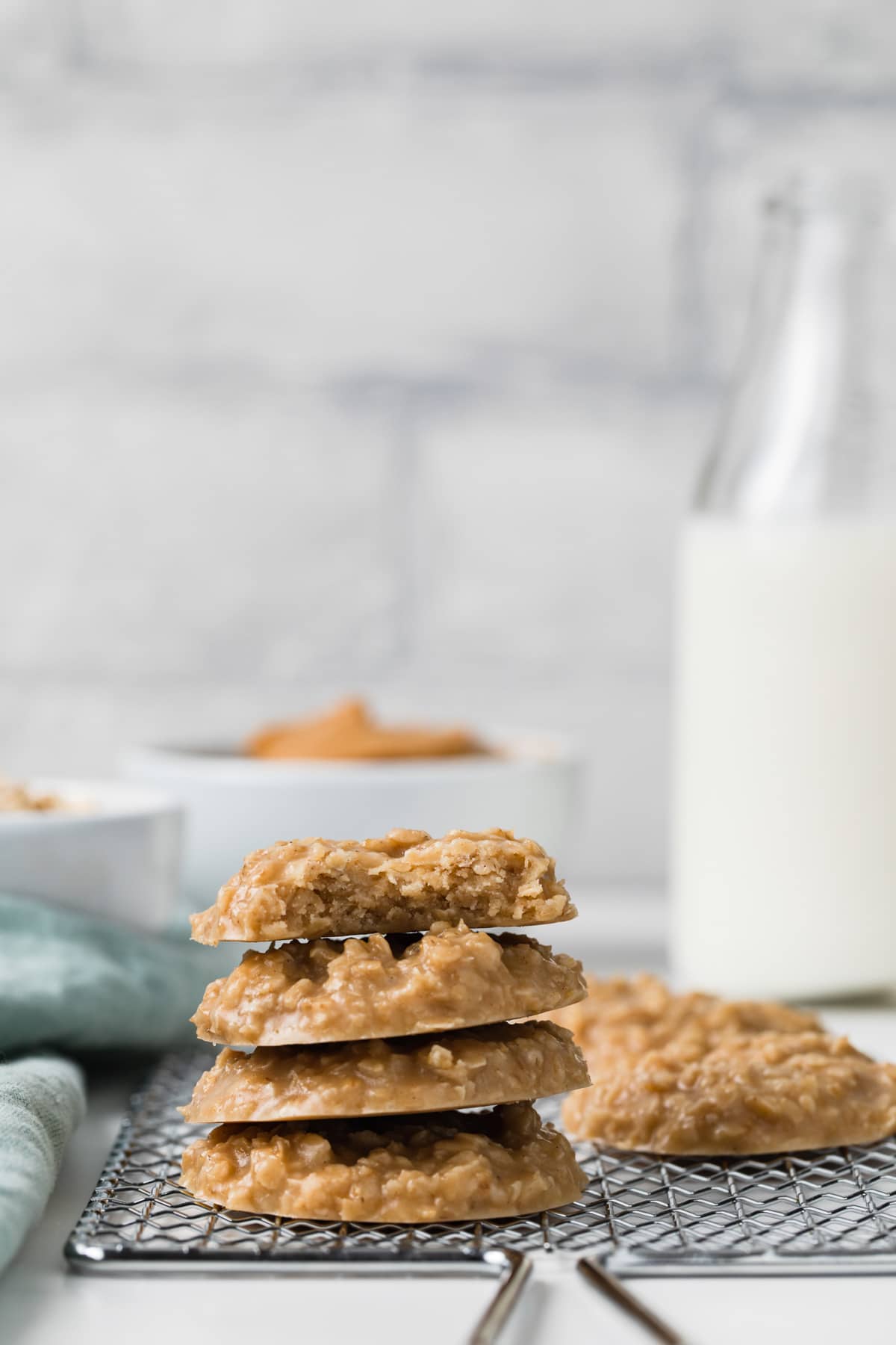 stack of four peanut butter no-bake cookies on a wire rack with a bowl of peanut butter and jar of milk behind it