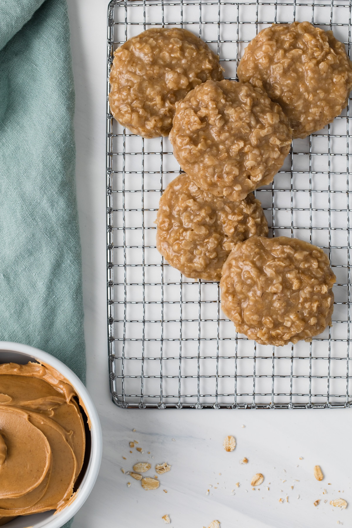 overhead view of peanut butter no bake cookies scattered on a wire rack next to a bowl of peanut butter and light blue-green cloth