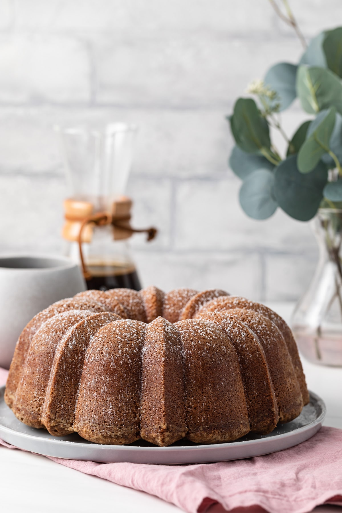 front view of whole pound cake on grey plate over a pink napkin with coffee and plant in background