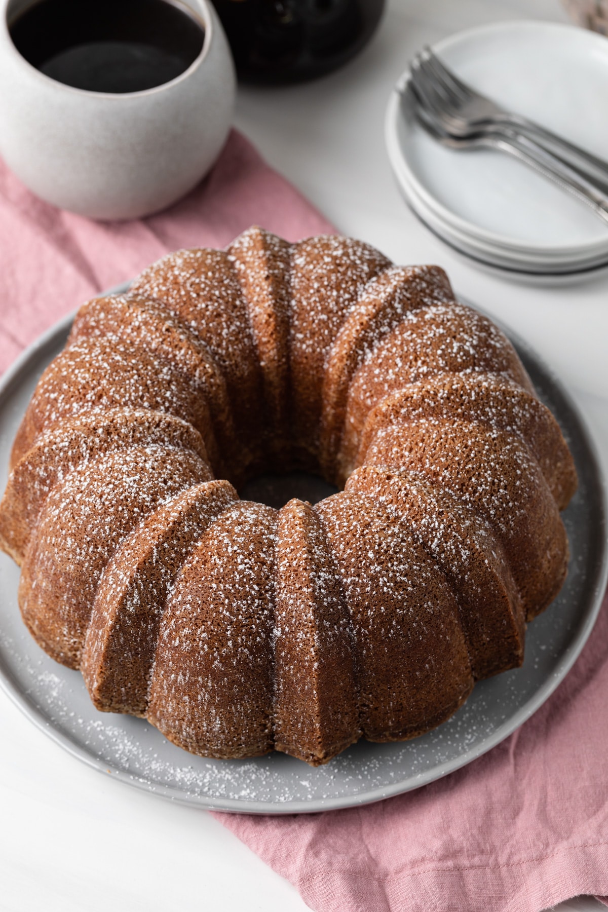 angled view of whole pound cake on grey plate with pink napkin and cup of coffee