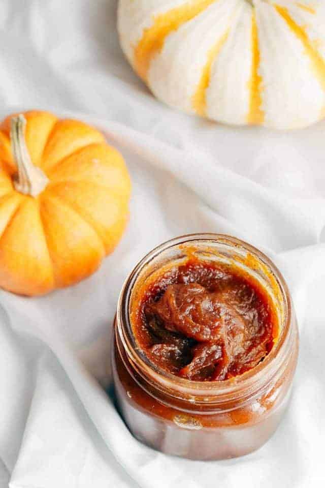 A glass jar full of pumpkin jam sitting on a light blue napkin with pumpkins in the back ground.