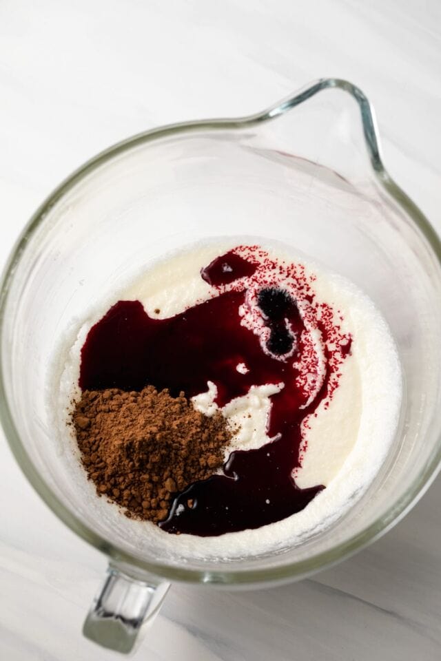 Red food coloring and cocoa powder added to sugar in glass bowl.