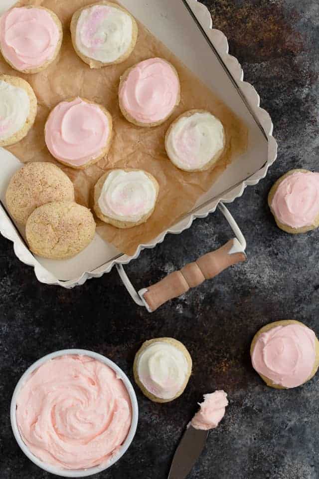 Frosted sour cream cookies next to a try filled with cookies and a white bowl filled with pink frosting.