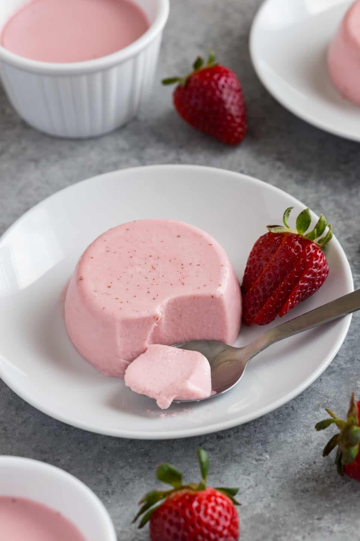 Creamy strawberry panna cotta on a white plate with a spoon taking a bite out.