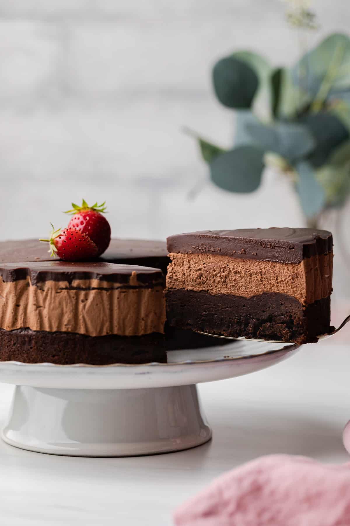 A close up view of triple chocolate mousse cake sitting on a white cake plate with large strawberries on top of the cake.
